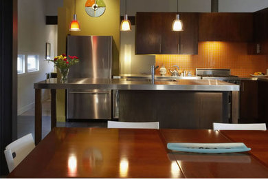 Example of a mid-sized trendy l-shaped eat-in kitchen design in Other with flat-panel cabinets, dark wood cabinets, stainless steel countertops, orange backsplash, glass tile backsplash, stainless steel appliances and an island