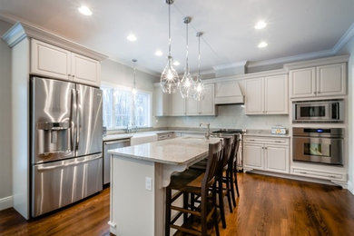 Inspiration for a large classic l-shaped kitchen/diner in New York with a belfast sink, raised-panel cabinets, white cabinets, granite worktops, white splashback, glass tiled splashback, stainless steel appliances, medium hardwood flooring and an island.