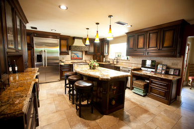 Example of a large tuscan u-shaped travertine floor eat-in kitchen design in Los Angeles with raised-panel cabinets, dark wood cabinets, granite countertops, beige backsplash, an island, a farmhouse sink, stone tile backsplash and stainless steel appliances