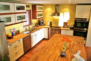 Inredning av ett klassiskt stort kök, med rostfria vitvaror, en köksö, en dubbel diskho, skåp i shakerstil, vita skåp, träbänkskiva, mellanmörkt trägolv och gult stänkskydd