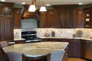 Cette image montre une cuisine en bois foncé avec un placard avec porte à panneau encastré, un plan de travail en granite, une crédence beige, une crédence en carrelage de pierre, un électroménager en acier inoxydable, un sol en bois brun et îlot.