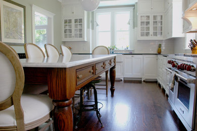 Example of a large transitional l-shaped dark wood floor and brown floor eat-in kitchen design in Chicago with a farmhouse sink, white cabinets, white backsplash, ceramic backsplash, stainless steel appliances, no island, shaker cabinets and solid surface countertops