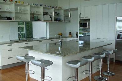 Example of a large minimalist l-shaped light wood floor and brown floor open concept kitchen design in Vancouver with a double-bowl sink, flat-panel cabinets, white cabinets, granite countertops, stainless steel appliances and an island