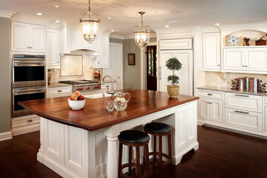 Open concept kitchen - large transitional l-shaped dark wood floor and brown floor open concept kitchen idea in Kansas City with a farmhouse sink, beaded inset cabinets, white cabinets, wood countertops, white backsplash, porcelain backsplash, paneled appliances and an island