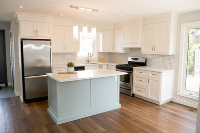 Inspiration for a mid-sized transitional l-shaped medium tone wood floor and brown floor open concept kitchen remodel in Toronto with a double-bowl sink, shaker cabinets, white cabinets, solid surface countertops, white backsplash, ceramic backsplash, stainless steel appliances and an island