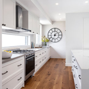 Design ideas for a large beach style l-shaped kitchen in Other with an undermount sink, recessed-panel cabinets, white cabinets, white splashback, panelled appliances, medium hardwood floors, with island, brown floor and grey benchtop.
