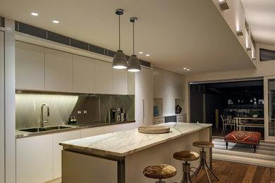 Modern inredning av ett mellanstort kök, med en nedsänkt diskho, släta luckor, vita skåp, bänkskiva i rostfritt stål, stänkskydd med metallisk yta, rostfria vitvaror, mellanmörkt trägolv och en köksö