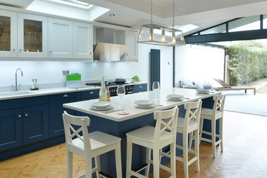 This is an example of a medium sized contemporary kitchen/diner in Cornwall with a built-in sink, shaker cabinets, blue cabinets, quartz worktops, glass sheet splashback, black appliances, light hardwood flooring, an island and white worktops.