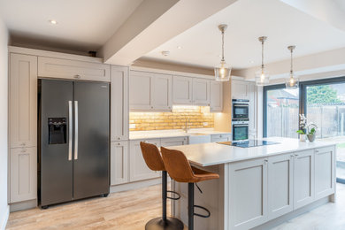 This is an example of a large traditional single-wall open plan kitchen in Hertfordshire with a belfast sink, shaker cabinets, beige cabinets, quartz worktops, brick splashback, stainless steel appliances, laminate floors, an island, white worktops, beige splashback and beige floors.