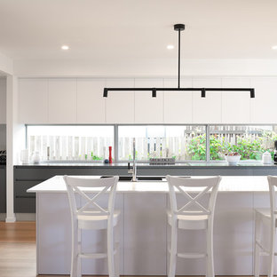 Photo of a beach style kitchen in Gold Coast - Tweed with a triple-bowl sink, flat-panel cabinets, white cabinets, window splashback, stainless steel appliances, light hardwood floors, with island, beige floor and grey benchtop.