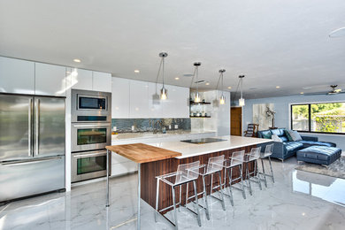 Example of a mid-sized trendy single-wall marble floor and gray floor open concept kitchen design in Miami with an undermount sink, flat-panel cabinets, white cabinets, solid surface countertops, metallic backsplash, metal backsplash, stainless steel appliances, an island and white countertops