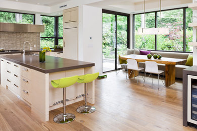 Example of a large trendy l-shaped medium tone wood floor and brown floor eat-in kitchen design in Vancouver with stainless steel appliances, flat-panel cabinets, white cabinets, quartz countertops, an undermount sink, beige backsplash, glass tile backsplash, an island and brown countertops
