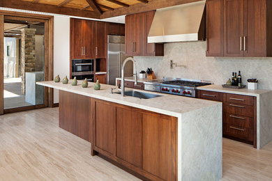 Modelo de cocina contemporánea con fregadero bajoencimera, armarios estilo shaker, puertas de armario de madera en tonos medios, salpicadero verde, electrodomésticos de acero inoxidable, una isla y suelo de travertino