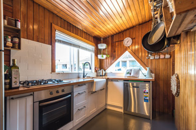 オークランドにあるラスティックスタイルのおしゃれなL型キッチン (エプロンフロントシンク、シェーカースタイル扉のキャビネット、白いキャビネット、木材カウンター、白いキッチンパネル、サブウェイタイルのキッチンパネル、シルバーの調理設備、コンクリートの床、アイランドなし、黒い床、茶色いキッチンカウンター、板張り天井) の写真
