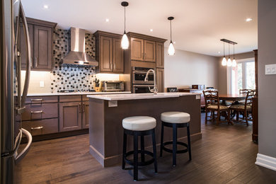 Large traditional l-shaped kitchen/diner in Toronto with a built-in sink, shaker cabinets, brown cabinets, engineered stone countertops, beige splashback, metro tiled splashback, stainless steel appliances, medium hardwood flooring, multiple islands and brown floors.