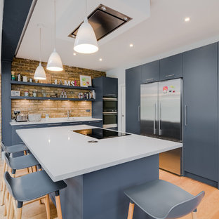 This is an example of a medium sized contemporary l-shaped kitchen in London with flat-panel cabinets, composite countertops, white splashback, stainless steel appliances, an island, a submerged sink, blue cabinets, brick splashback, medium hardwood flooring, brown floors and white worktops.