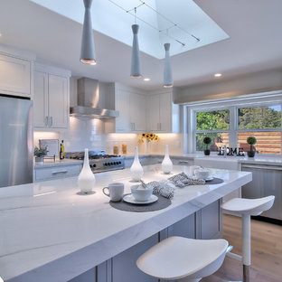 Photo of a modern l-shaped eat-in kitchen in San Francisco with a farmhouse sink, shaker cabinets, white cabinets, quartzite benchtops, white splashback, porcelain splashback, stainless steel appliances, light hardwood floors, with island and green floor.