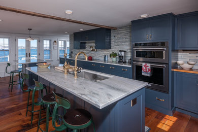 Design ideas for a large coastal galley kitchen/diner in Boston with a belfast sink, recessed-panel cabinets, blue cabinets, engineered stone countertops, multi-coloured splashback, glass tiled splashback, stainless steel appliances, medium hardwood flooring, multiple islands, brown floors and white worktops.