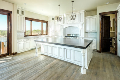 Large rustic l-shaped open plan kitchen in Denver with a belfast sink, recessed-panel cabinets, white cabinets, granite worktops, grey splashback, matchstick tiled splashback, stainless steel appliances, dark hardwood flooring and an island.