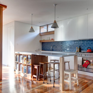 Contemporary galley open plan kitchen in Sydney with flat-panel cabinets, white cabinets, blue splashback, stainless steel appliances, medium hardwood floors, with island, brown floor and grey benchtop.