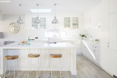 Photo of a traditional l-shaped kitchen in Sydney with a submerged sink, shaker cabinets, white cabinets, white splashback, integrated appliances, light hardwood flooring, an island, beige floors and white worktops.