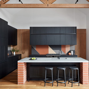 This is an example of a large industrial l-shaped kitchen in Sydney with black cabinets, marble benchtops, cement tile splashback, black appliances, medium hardwood floors, with island, black benchtop, exposed beam, vaulted, an undermount sink, flat-panel cabinets, multi-coloured splashback and brown floor.
