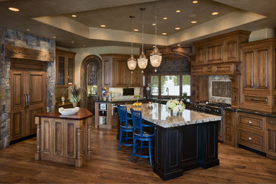 Large arts and crafts u-shaped medium tone wood floor and brown floor open concept kitchen photo in Other with a farmhouse sink, raised-panel cabinets, medium tone wood cabinets, granite countertops, gray backsplash, ceramic backsplash, paneled appliances and two islands