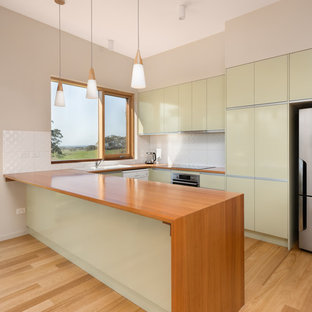 Design ideas for a mid-sized contemporary l-shaped eat-in kitchen in Other with green cabinets, wood benchtops, white splashback, ceramic splashback, light hardwood floors, a drop-in sink, flat-panel cabinets, stainless steel appliances, a peninsula, beige floor and brown benchtop.