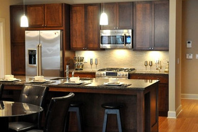 Example of a mid-sized transitional single-wall light wood floor and brown floor eat-in kitchen design in Indianapolis with a drop-in sink, flat-panel cabinets, dark wood cabinets, granite countertops, beige backsplash, stone tile backsplash, stainless steel appliances and an island