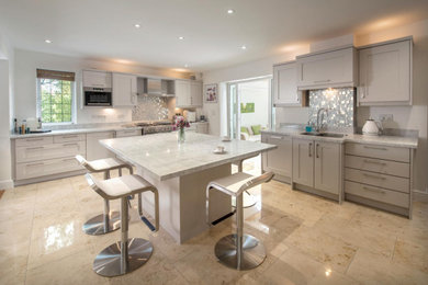 This is an example of a large contemporary kitchen/diner in Kent with shaker cabinets, a submerged sink, beige cabinets, engineered stone countertops, metallic splashback, mosaic tiled splashback, stainless steel appliances, travertine flooring, an island, beige floors and white worktops.