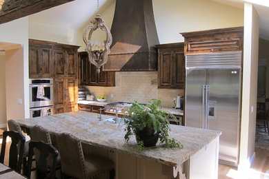 Mid-sized elegant l-shaped light wood floor and brown floor open concept kitchen photo in San Francisco with a farmhouse sink, dark wood cabinets, granite countertops, beige backsplash, stainless steel appliances, an island, raised-panel cabinets, travertine backsplash and multicolored countertops