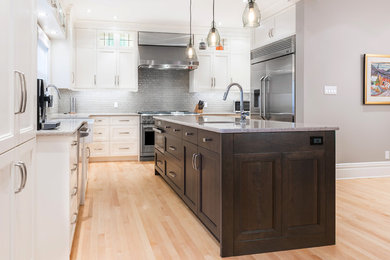 Example of a transitional light wood floor kitchen design in Ottawa with a farmhouse sink, recessed-panel cabinets, dark wood cabinets, quartz countertops, gray backsplash, glass tile backsplash, stainless steel appliances and an island