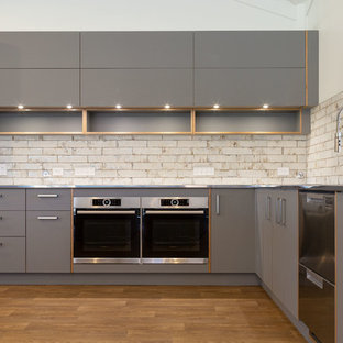 Large industrial l-shaped eat-in kitchen in Other with grey cabinets, multiple islands, a drop-in sink, flat-panel cabinets, laminate benchtops, white splashback, brick splashback, stainless steel appliances, dark hardwood floors, brown floor and grey benchtop.