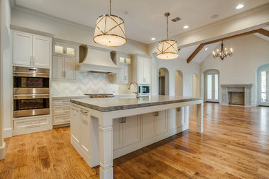 Large transitional single-wall medium tone wood floor open concept kitchen photo in Dallas with a farmhouse sink, white backsplash, stainless steel appliances, beaded inset cabinets, beige cabinets, marble countertops, stone tile backsplash and an island
