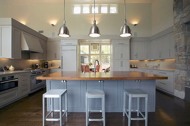 Example of a mid-sized arts and crafts u-shaped dark wood floor eat-in kitchen design in New York with a farmhouse sink, shaker cabinets, gray cabinets, wood countertops, beige backsplash, paneled appliances and an island