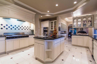 Example of a large trendy u-shaped ceramic tile eat-in kitchen design in Los Angeles with a drop-in sink, raised-panel cabinets, white cabinets, granite countertops, white backsplash, porcelain backsplash, stainless steel appliances and an island