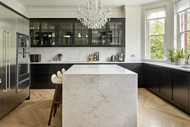 This is an example of a classic u-shaped kitchen in London with a submerged sink, shaker cabinets, black cabinets, white splashback, stone slab splashback, stainless steel appliances, light hardwood flooring, an island, beige floors and white worktops.