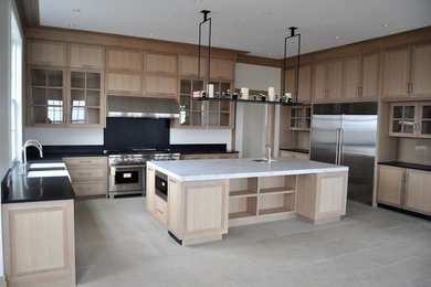 This is an example of a large classic u-shaped enclosed kitchen in Boston with limestone flooring, an island, a belfast sink, shaker cabinets, light wood cabinets, composite countertops, black splashback, stone slab splashback, stainless steel appliances and beige floors.