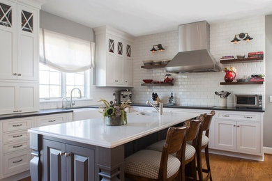 Eat-in kitchen - large transitional l-shaped medium tone wood floor and brown floor eat-in kitchen idea in New York with a farmhouse sink, recessed-panel cabinets, white cabinets, solid surface countertops, white backsplash, subway tile backsplash, stainless steel appliances, an island and black countertops