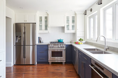 Inspiration for a mid-sized transitional l-shaped medium tone wood floor and brown floor kitchen remodel in Toronto with a double-bowl sink, shaker cabinets, blue cabinets, solid surface countertops, white backsplash, ceramic backsplash, stainless steel appliances and no island