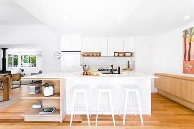На фото: параллельная кухня-гостиная в современном стиле с врезной мойкой, плоскими фасадами, белыми фасадами, белым фартуком, светлым паркетным полом, островом, бежевым полом и белой столешницей