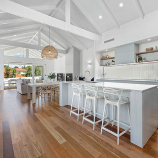 Mid-sized beach style galley open plan kitchen in Brisbane with an undermount sink, flat-panel cabinets, window splashback, black appliances, with island, brown floor, exposed beam, vaulted, grey cabinets, medium hardwood floors and white benchtop.