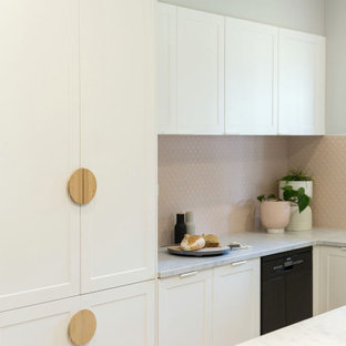 Réalisation d'une petit cuisine américaine nordique en L avec un évier encastré, un placard avec porte à panneau encastré, des portes de placard blanches, un plan de travail en quartz modifié, une crédence rose, une crédence en mosaïque, un électroménager noir, un sol en bois brun, un îlot central et un plan de travail blanc.