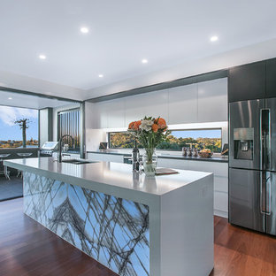 Large contemporary single-wall open plan kitchen in Brisbane with a double-bowl sink, flat-panel cabinets, white cabinets, black appliances, medium hardwood floors, brown floor, white benchtop, quartz benchtops, white splashback, glass sheet splashback and with island.
