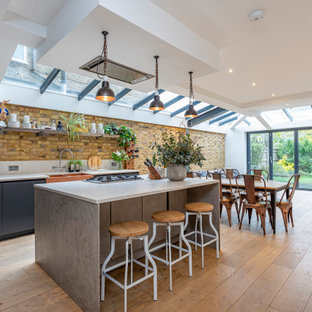 Contemporary galley kitchen in Sussex with a belfast sink, flat-panel cabinets, grey cabinets, brown splashback, brick splashback, stainless steel appliances, medium hardwood flooring, an island, brown floors and white worktops.