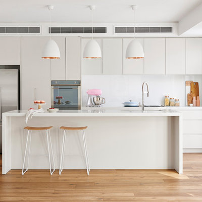 Example of a trendy single-wall light wood floor and beige floor open concept kitchen design in Melbourne with an undermount sink, flat-panel cabinets, beige cabinets, white backsplash, stainless steel appliances, an island, quartz countertops and glass sheet backsplash