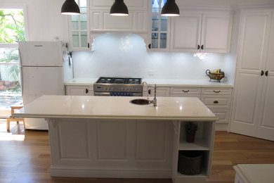 Klassisk inredning av ett stort kök, med en rustik diskho, luckor med upphöjd panel, vita skåp, bänkskiva i kvarts, vitt stänkskydd, stänkskydd i keramik, rostfria vitvaror, ljust trägolv och en köksö