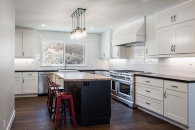Elegant l-shaped medium tone wood floor and brown floor eat-in kitchen photo in Salt Lake City with a farmhouse sink, recessed-panel cabinets, white cabinets, white backsplash, ceramic backsplash, stainless steel appliances and an island