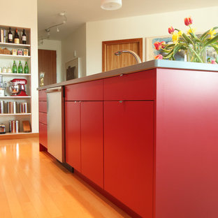 Modelo de cocina moderna, pequeña, abierta, con fregadero bajoencimera, armarios con paneles lisos, puertas de armario rojas, encimera de acrílico, salpicadero de metal, electrodomésticos de acero inoxidable, suelo de madera en tonos medios y una isla