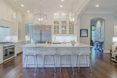 Elegant l-shaped medium tone wood floor open concept kitchen photo in Dallas with an island, a farmhouse sink, recessed-panel cabinets, white cabinets, marble countertops, white backsplash, subway tile backsplash and stainless steel appliances